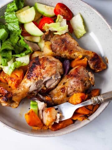 A large plate filled with three roasted chicken drumsticks, sweet potatoes, and a green salad with romaine lettuce, cucumbers, and cherry tomatoes.