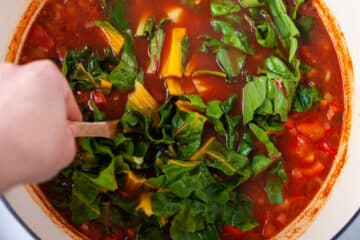 A hand stirring chopped rainbow chard in a red tomato based vegetable soup.