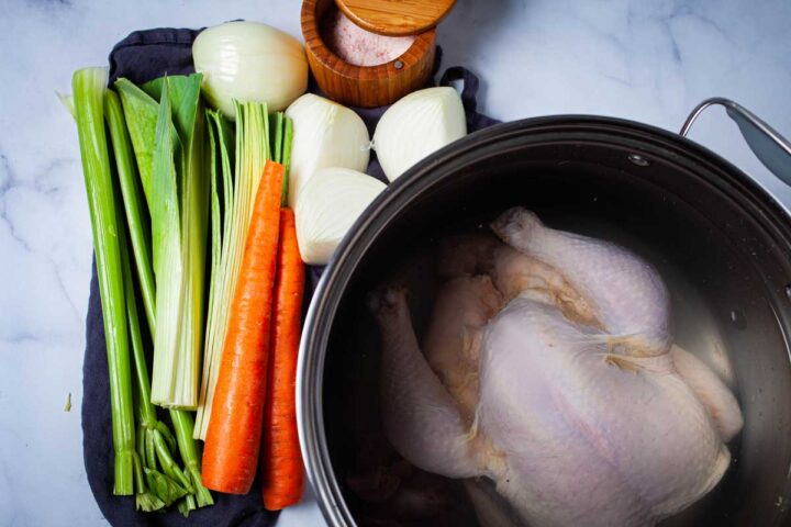 A large pot containing a whole raw chicken covered with water, next to fresh chopped vegetables like celery, leek, carrots, onions, and a pot of salt.