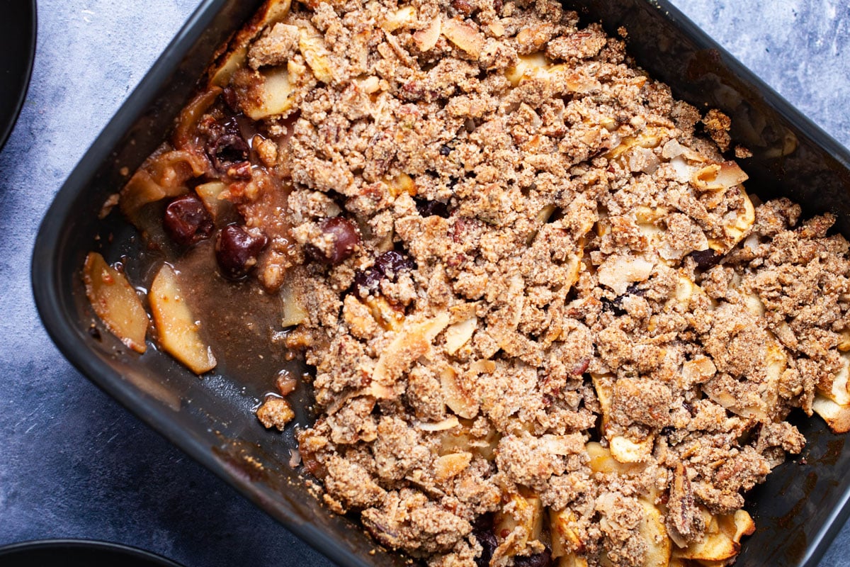 A spoon taken out of a baking pan containing baked apple and cherry crisp without oats.