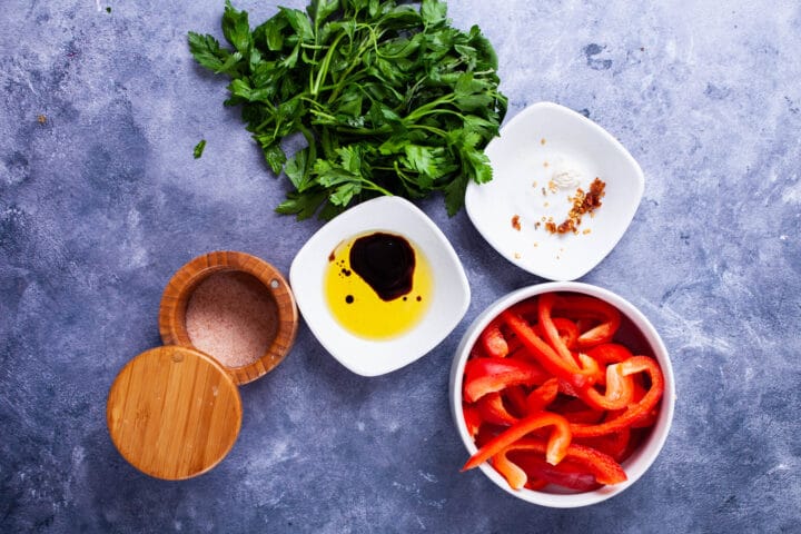 Slices of red bell pepper, salt, olive oil, balsamic vinegar, chili pepper flakes, and fresh parsley leaves displayed on a table.