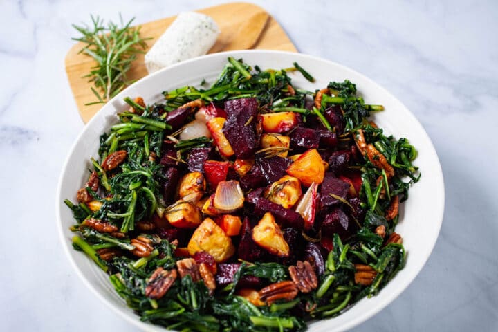 A large plate filled with roasted beets, potatoes, sauteed dandelion greens, pecans, next to a wooden plate topped with extra goat cheese and Rosemary.