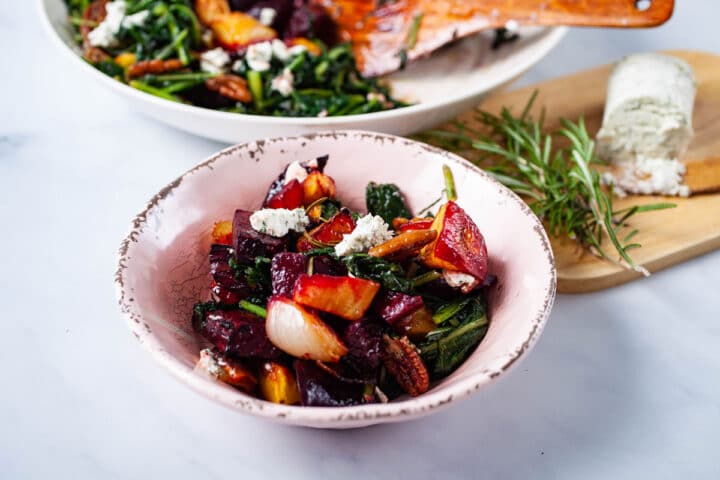 A small plate filled with roasted beets, potatoes, sauteed dandelion greens, pecans, topped with goat cheese, with a wooden plate with extra goat cheese and Rosemary in the background.