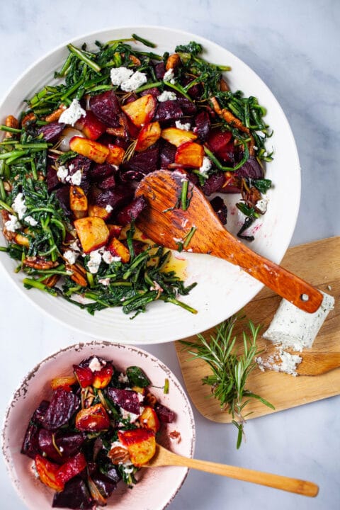 A large plate filled with roasted beets, potatoes, sauteed dandelion greens, pecans, topped with goat cheese, and next to a wooden plate with extra goat cheese and Rosemary.