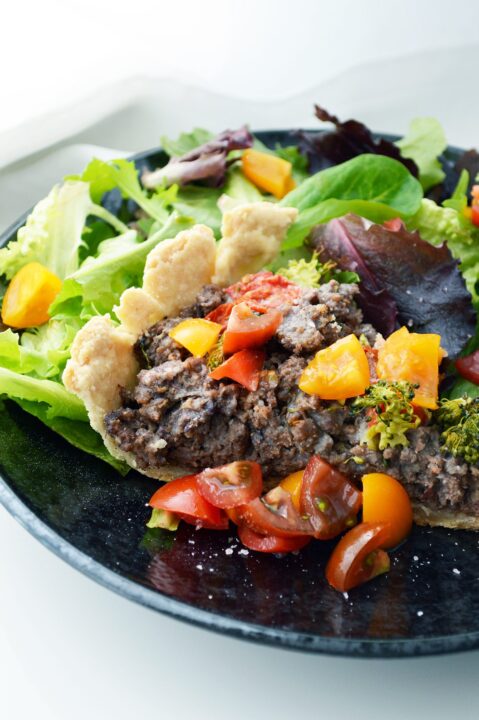 rustic bison skillet slice beside a bed of salad and garnished with fresh sliced cherry tomatoes on a black plate.