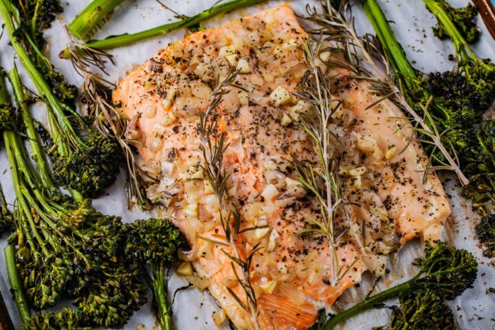 Baked salmon topped with buttery onions and surrounded by baby broccoli on a baking sheet.