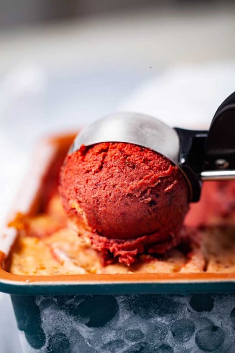 A bowl of berry nice cream in an ice cream scoop resting on top of more ice cream in a container.