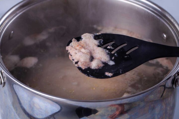 A large spoon resting over a pot with protein scum from poaching a chicken.
