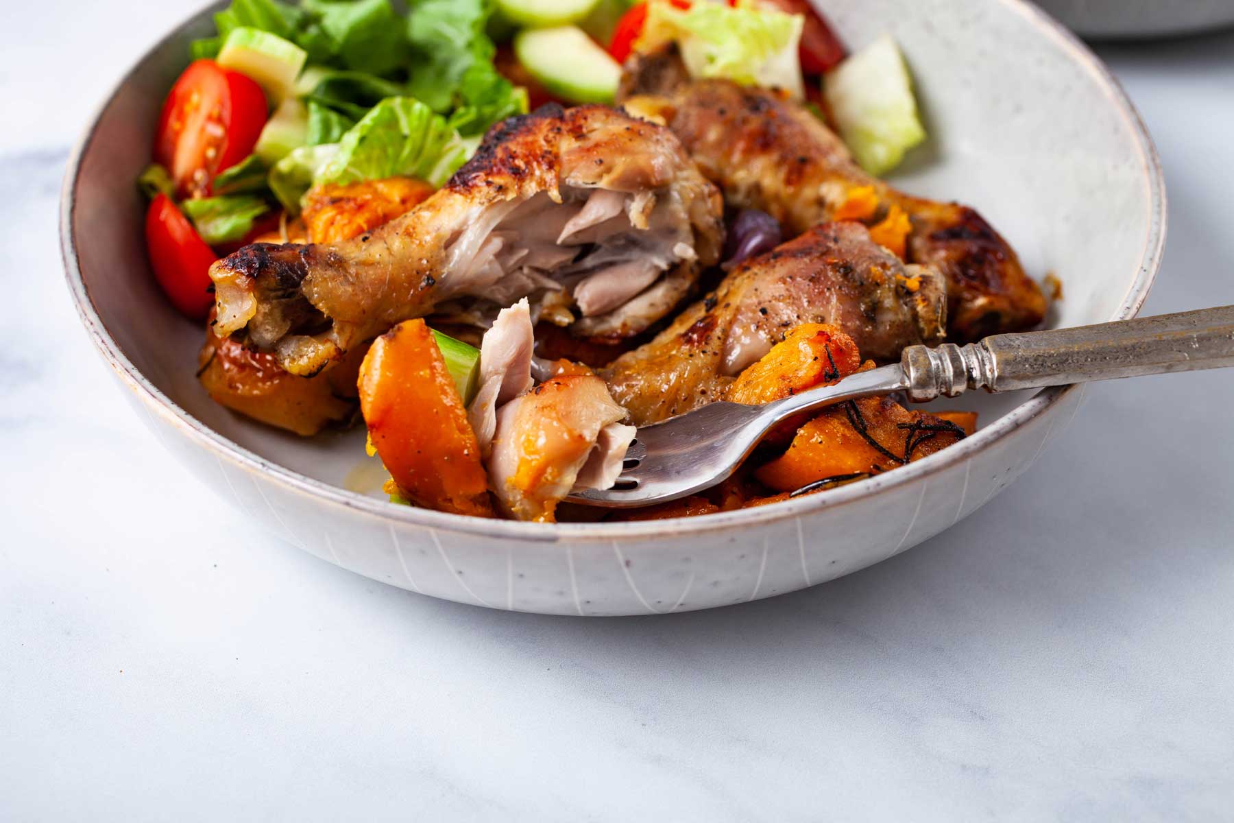 A fork resting on a plate filled with juice chicken drumsticks, roasted sweet potatoes and apples.