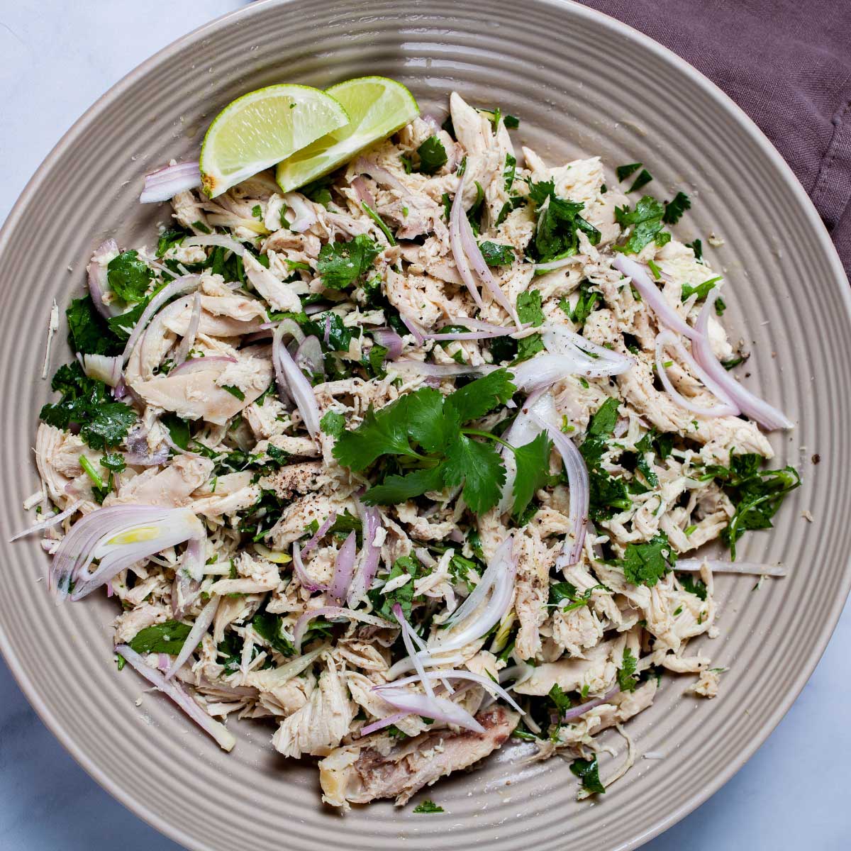 A large deep plate filled with shredded chicken salad with cilantro, olive oil, shallots, salt and pepper, and lime wedges on the side.