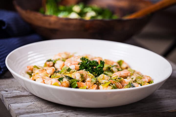 A large white plate filled with sauteed shrimp and leeks, placed on a wooden table.