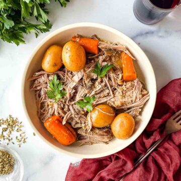 Pork roast in a bowl with small potatoes and carrots, topped with parsley leaves.