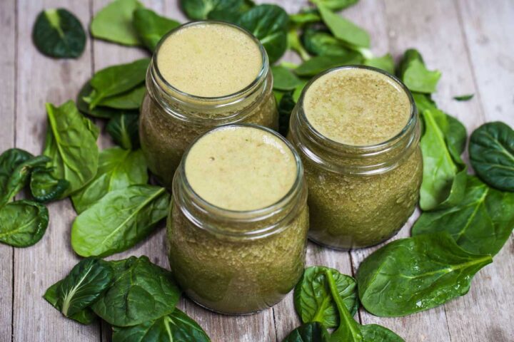 Three small glass jars filled with green smoothie and surrounded by fresh spinach leaves.