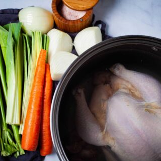 A large pot containing a whole raw chicken covered with water, next to fresh chopped vegetables like celery, leek, carrots, onions, and a pot of salt.