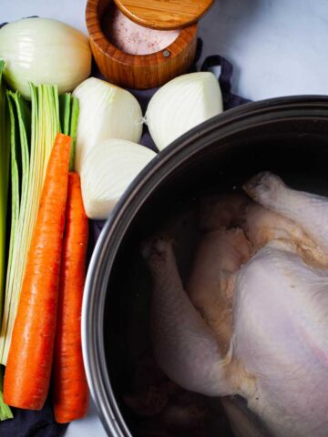 A large pot containing a whole raw chicken covered with water, next to fresh chopped vegetables like celery, leek, carrots, onions, and a pot of salt.