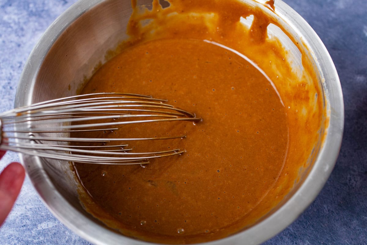 A whisk resting in a stainless steel bowl containing a brown batter.