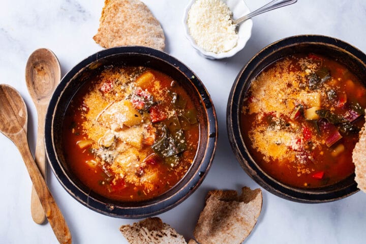 A tomato based vegetable soup with potatoes served in a wooden bowls, topped with grated Parmesan cheese and served with pieces of bread on the side.