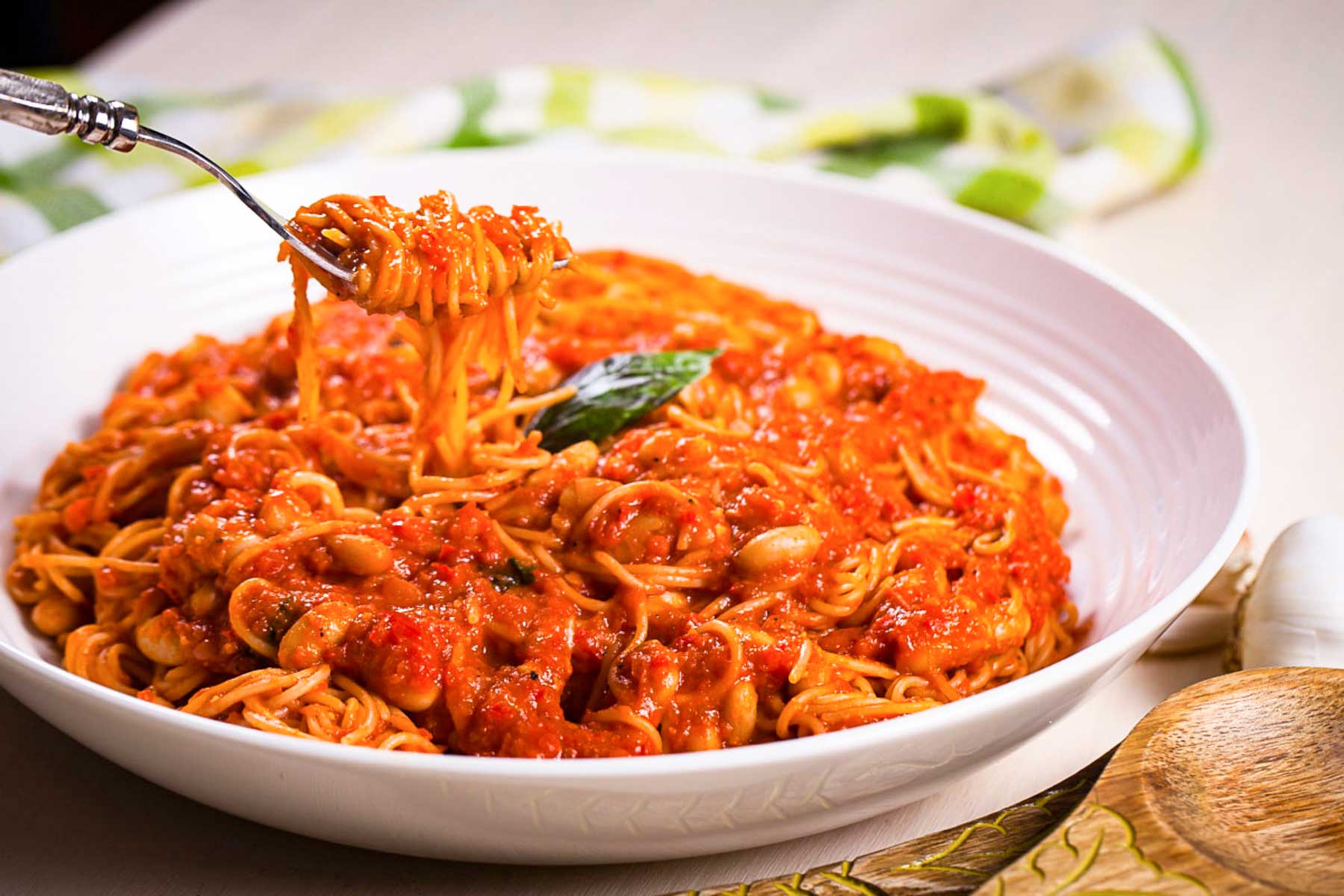 A fork lifting a bite out of a plate with spaghetti noodles drenched in a red tomato sauce with beans.