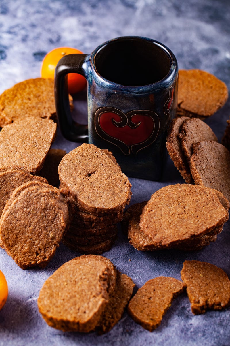 Stacked brown and thin Speculaas cookies surrounding a handmade mug with a heart shape on it.