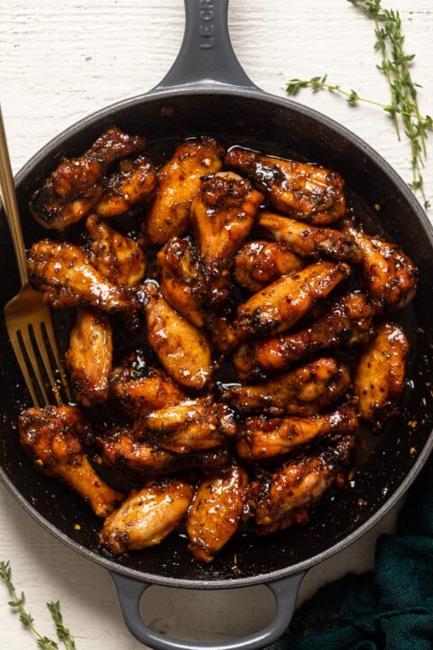 an overhead shot of sticky peach chicken wings in a enameled cast iron skillet with a golden fork tucked in the side and fresh thyme on the white surface beside the skillet.
