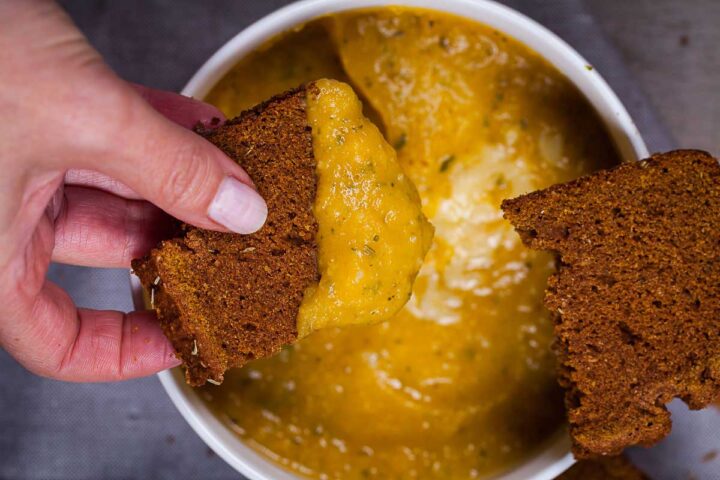 A women's hand holding a piece of farmer's bread and dipping it in a bowl of yellow squash soup.
