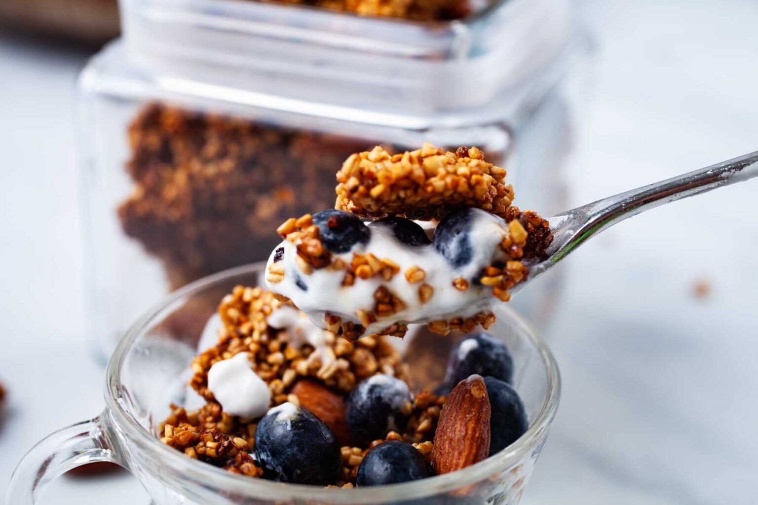 A spoonful of granola with yogurt and blueberries is being lifted from a glass bowl. The bowl is filled with granola, almonds, yogurt, and fresh blueberries, while a glass container of granola sits in the background.