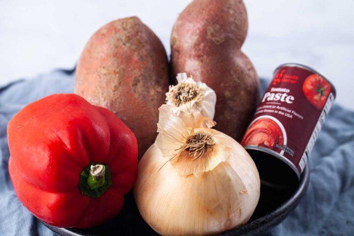 A bowl filled with one red bell pepper, one yellow onion, two sweet potatoes, and one can of tomato paste.