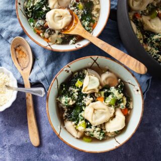 Two bowls filled with Straciatella soup with tortellini next to a large pot of soup, extra Parmesan cheese and wooden spoons on the side.