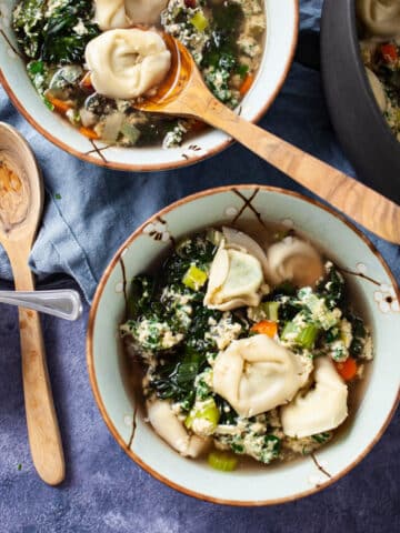 Two bowls filled with Straciatella soup with tortellini next to a large pot of soup, extra Parmesan cheese and wooden spoons on the side.