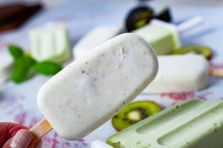 A hand holding up a white-colored ice pop with other white and green-colored ice pops in the background.