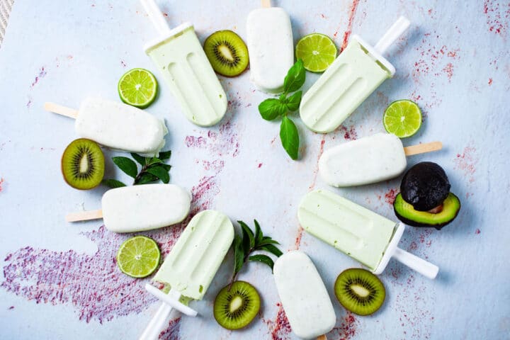 Green and white popsicles displayed in a circle on a table and decorated with slices of kiwi, lime, avocado, and mint leaves.