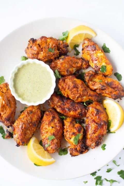 an overhead shot of tandoori chicken wings on a white plate with lemon wedges tucked amongst the wings and a small bowl of dipping sauce on the plate.