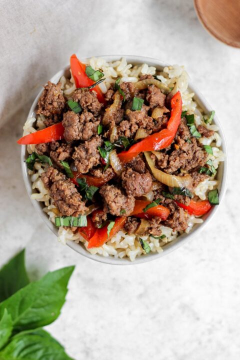 an overhead shot of ground bison thai basil stir fry in a white bowl with some thai basil on the table beside the bowl.