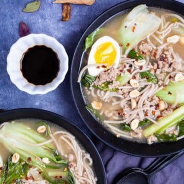 Two plates of bock choy soup with pork and noodles and garnished with chili peppers, peanuts, and hard-boiled egg.