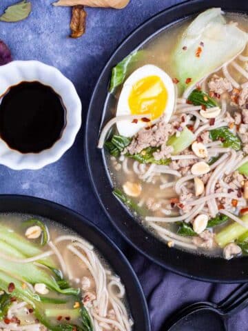 Two plates of bock choy soup with pork and noodles and garnished with chili peppers, peanuts, and hard-boiled egg.