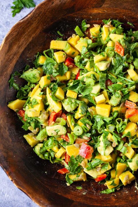 A large wooden bowl filled with fresh and creamy mango avocado salad, onions, cilantro, sunflower seeds and red bell peppers.
