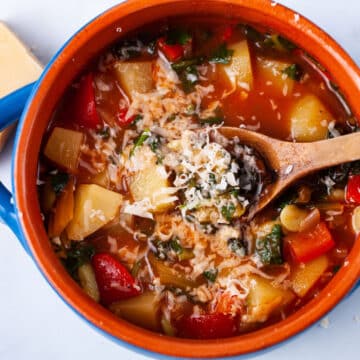 A spoon resting in a red tomato based vegetable soup topped with parmesan cheese.