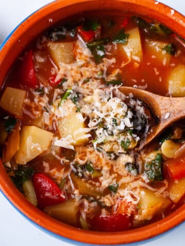 A spoon resting in a red tomato based vegetable soup topped with parmesan cheese.