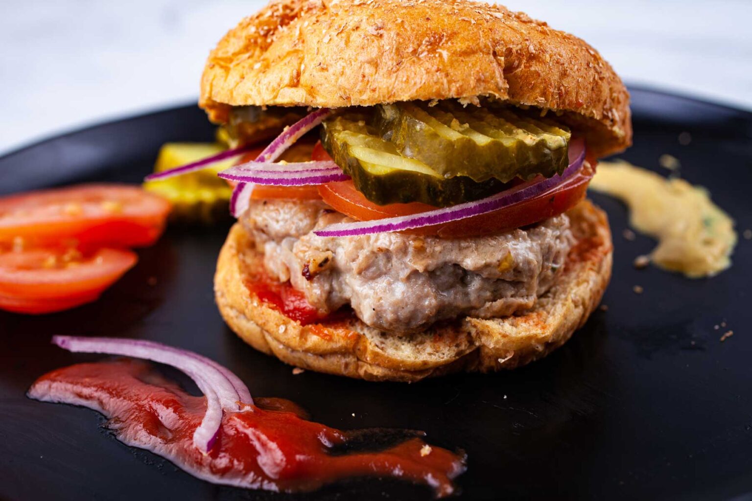 A close-up of a chicken burger on a black plate. The burger includes a sesame seeded bun, pickles, a tomato slice, red onion rings, and chicken patty. A smear of ketchup and mustard is on the side, with tomato slices as garnish.