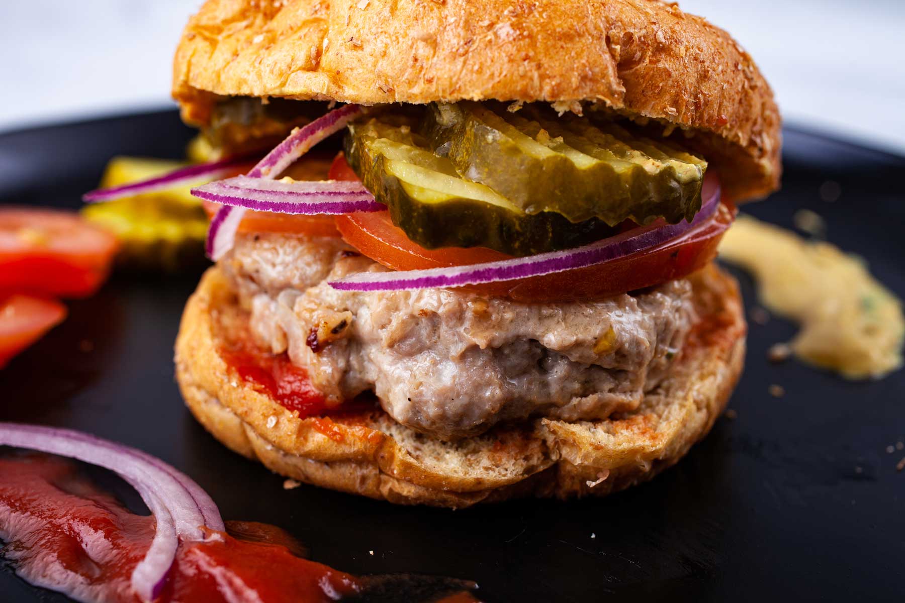 Gluten free turkey burger topped with slices of tomato, thinly sliced red onions, pickles in a bun next to a dollop of ketchup.