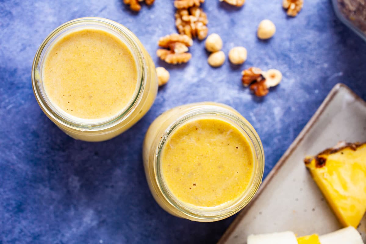 Two glasses filled with creamy yellow smoothie next to pineapple and nut snacks.