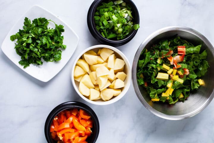 Chopped green onions, chopped parsley leaves, chopped rainbow chard, chopped bell pepper, and cubed potatoes displayed on a table.