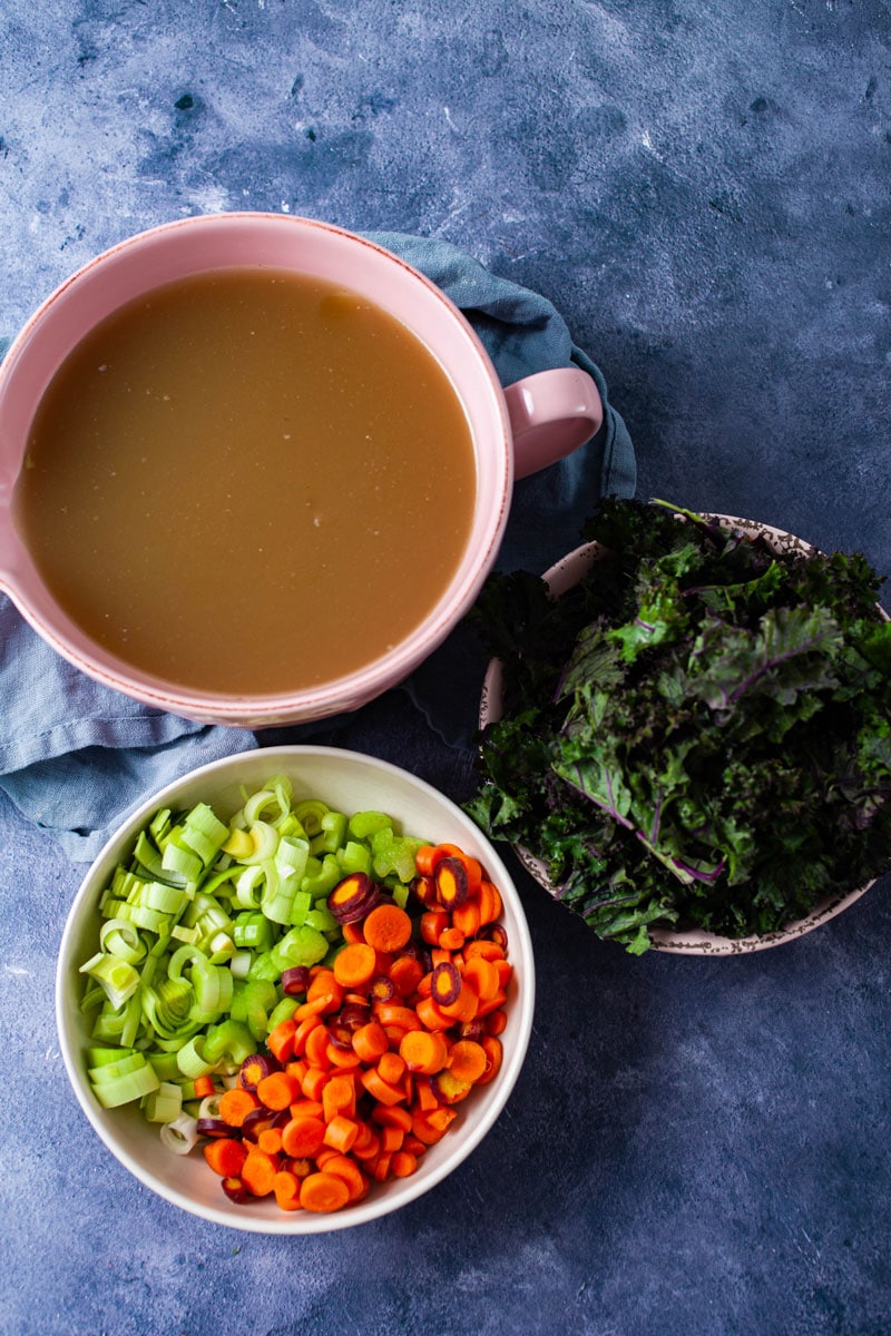 A large bowl with chicken stock, another bowl with chopped fresh veggies like carrots, leeks, and celery, and another bowl with chopped kale on a table.