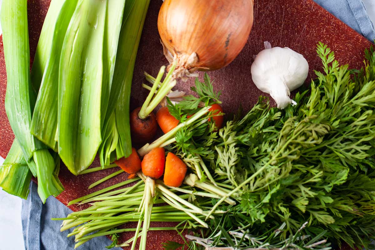 A yellow onion, carrot scraps, carrot greens, garlic, and leek leaves scattered on a cutting board.