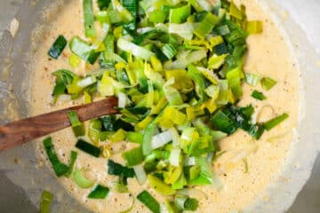 A wooden spoon stirring chopped leek in a batter in a bowl.