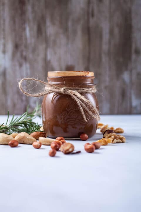 An opened jar with chocolate nut butter and surrounded by toasted nuts and Rosemary leaves.