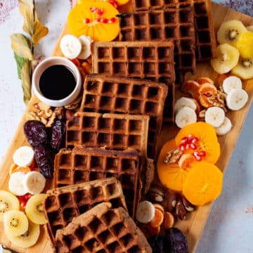 Golden brown waffles , an assortment of fresh and dried fruits, nuts, and a small syrup pourer beautifully arranged on a wooden board.