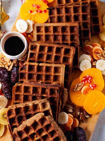 Golden brown waffles , an assortment of fresh and dried fruits, nuts, and a small syrup pourer beautifully arranged on a wooden board.