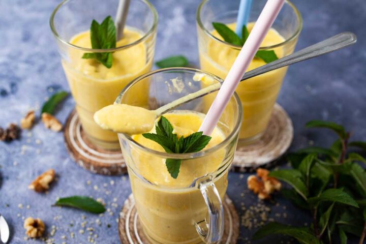 Three glasses filled with mango lassi, topped with mint leaves, and carrying straws. One has a large lassi filled teaspoon resting on top.