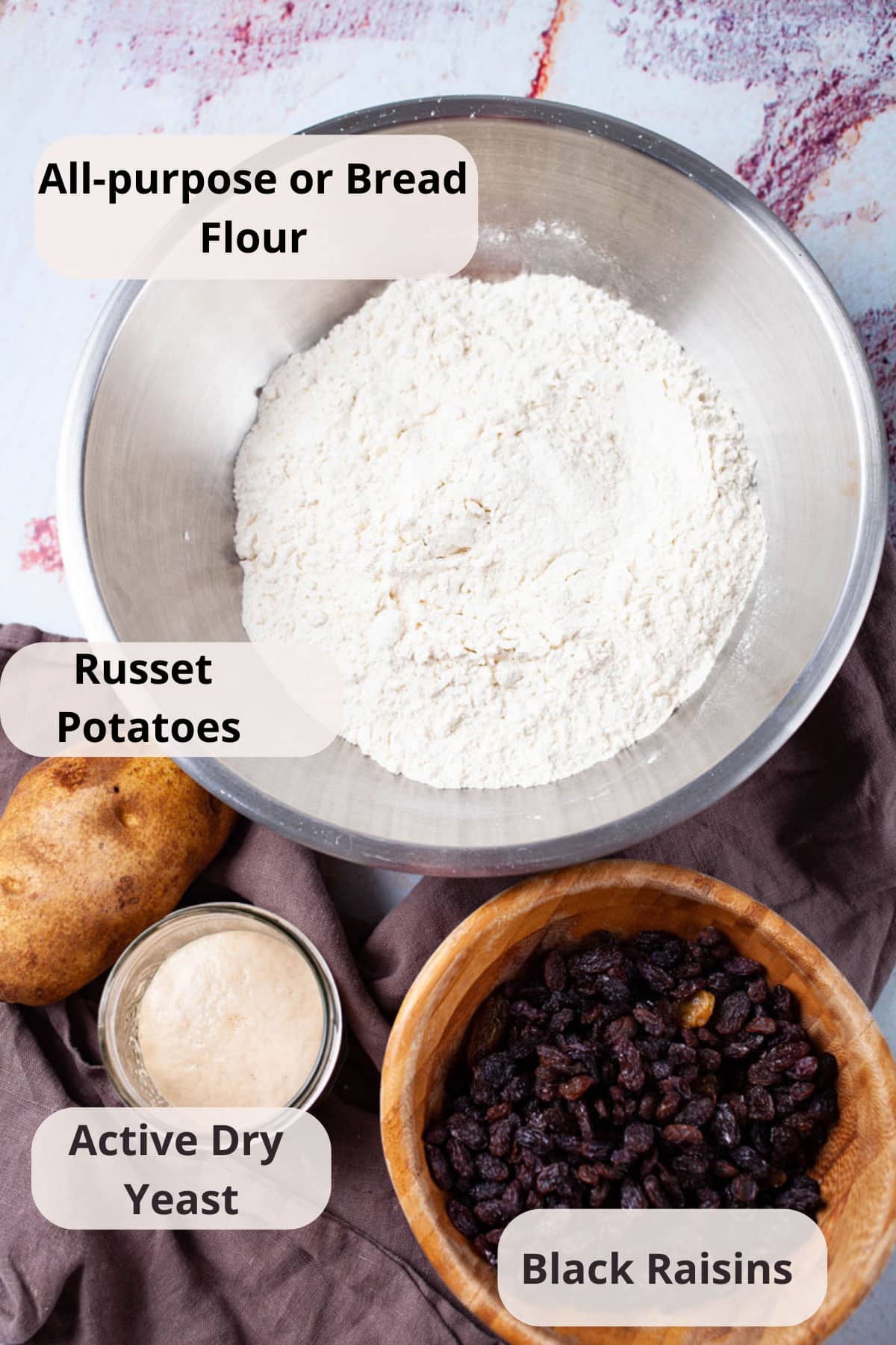 Ingredients such as a bowl of flour, a russet potato, leavened yeast, and a bowl of raisins displayed on a table.